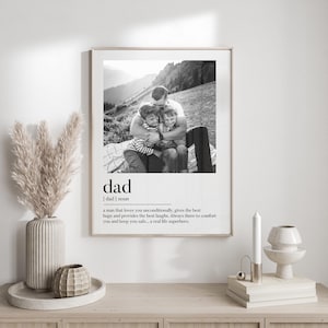 Peut inclure: Impression en noir et blanc encadr&eacute;e avec une photo d'un p&egrave;re &eacute;treignant deux enfants. Le mot "dad" et sa d&eacute;finition sont imprim&eacute;s en dessous. Le cadre est pos&eacute; sur une surface en bois clair.
