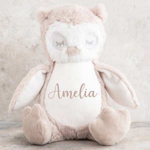 May include: A plush brown owl toy with white accents and closed eyes. The owl has a white patch on its chest with the name "Amelia" embroidered in pink.