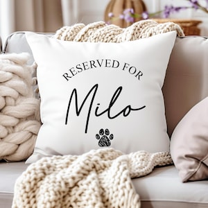 Puede incluir: Coj&iacute;n cuadrado blanco con el texto "RESERVED FOR Milo" en escritura negra. Una huella de pata negra est&aacute; debajo del nombre. El coj&iacute;n est&aacute; sobre un sof&aacute; gris con una manta de punto color crema.