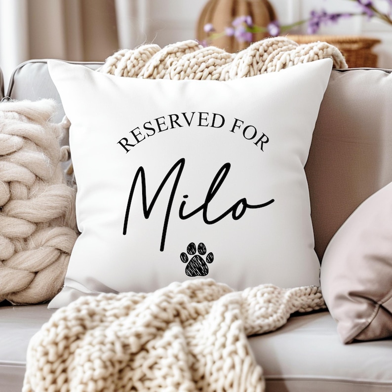 Puede incluir: Coj&iacute;n cuadrado blanco con el texto "RESERVED FOR Milo" en escritura negra. Una huella de pata negra est&aacute; debajo del nombre. El coj&iacute;n est&aacute; sobre un sof&aacute; gris con una manta de punto color crema.