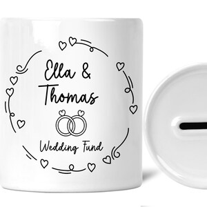 May include: White ceramic piggy bank with a slot on the top. The front of the bank has a black design with the text "Ella & Thomas Wedding Fund" and a ring design.
