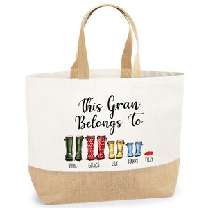 May include: A white canvas tote bag with brown jute handles and a brown jute bottom. The bag has a black graphic of a line of colorful rain boots with the text "This Gran Belongs To" above the boots and the names "Phil, Grace, Lily, Harry, Tilly" below the boots.