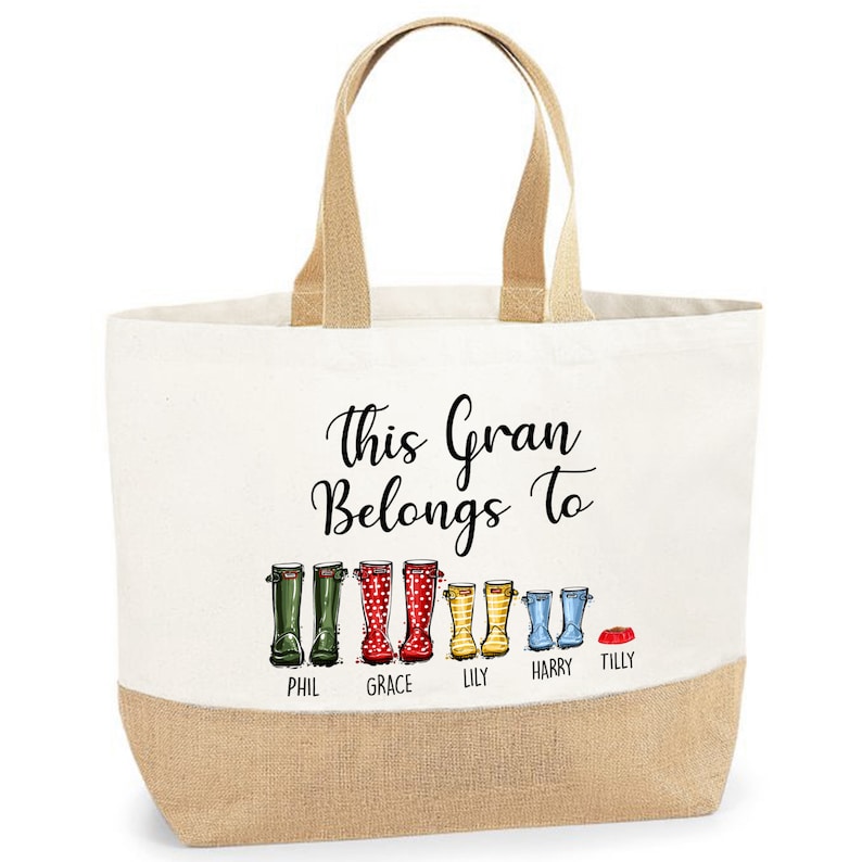 May include: A white canvas tote bag with brown jute handles and a brown jute bottom. The bag has a black graphic of a line of colorful rain boots with the text "This Gran Belongs To" above the boots and the names "Phil, Grace, Lily, Harry, Tilly" below the boots.