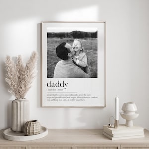 K&ouml;nnte beinhalten: Gerahmter Schwarz-Wei&szlig;-Druck mit einem Foto von Vater und Kind und dem Wort &bdquo;Daddy&ldquo; mit einer Definition. Der Druck wird auf einem Holzschrank angezeigt. Inneneinrichtung.