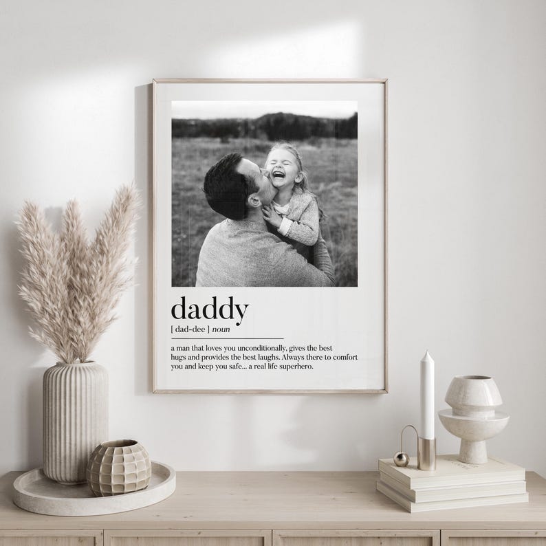 K&ouml;nnte beinhalten: Gerahmter Schwarz-Wei&szlig;-Druck mit einem Foto von Vater und Kind und dem Wort &bdquo;Daddy&ldquo; mit einer Definition. Der Druck wird auf einem Holzschrank angezeigt. Inneneinrichtung.