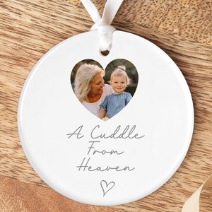 Puede incluir: Adorno de cer&aacute;mica blanca con un recorte de foto en forma de coraz&oacute;n de una mujer mayor y una ni&ntilde;a. El texto "A Cuddle From Heaven" est&aacute; escrito debajo de la foto con un peque&ntilde;o coraz&oacute;n en la parte inferior.