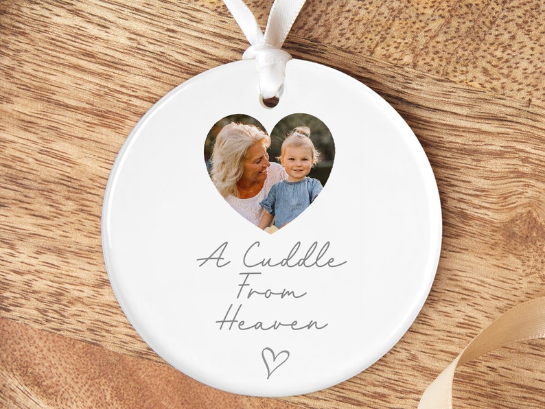 Puede incluir: Adorno de cer&aacute;mica blanca con un recorte de foto en forma de coraz&oacute;n de una mujer mayor y una ni&ntilde;a. El texto "A Cuddle From Heaven" est&aacute; escrito debajo de la foto con un peque&ntilde;o coraz&oacute;n en la parte inferior.