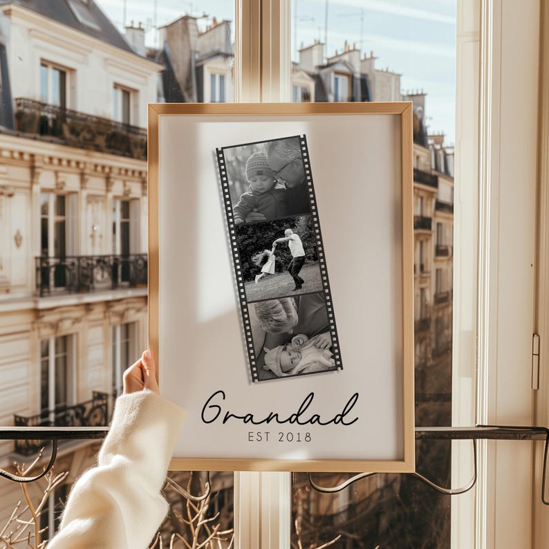 May include: A framed print featuring a black and white film strip of three family photos. The print is in a light wood frame, with the word "Grandad" and the year "EST 2018" in a script font at the bottom. The background shows a city view.