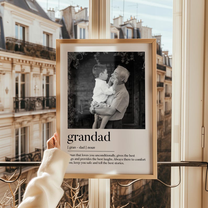May include: A framed black and white photograph featuring a grandfather holding a child. The frame is a light wood color. Below the photo, the word "grandad" is printed in a bold, sans-serif font, along with a definition.