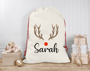 Puede incluir: Un saco de Santa de lona blanca con cordones de extracción rojos, que presenta una silueta de reno marrón con cuernos y una nariz roja. El saco está personalizado con el nombre "Sarah".