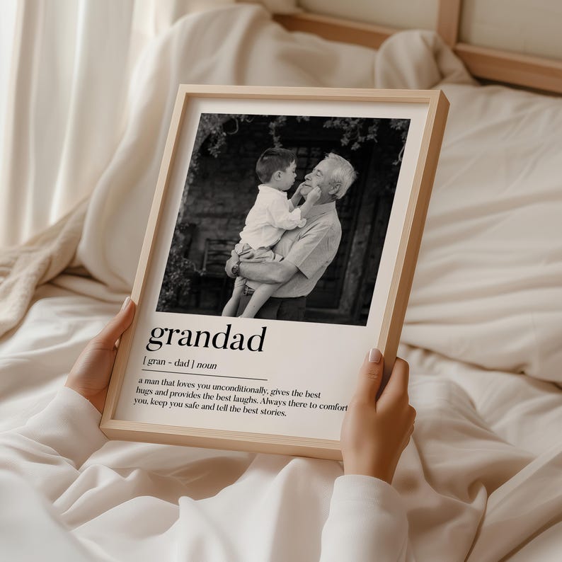 May include: A framed black and white photograph featuring a grandfather holding a child. The frame is light-colored wood. The word "grandad" is printed below the photo, along with a definition.