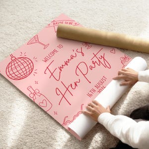 May include: A pink poster with red illustrations and text that reads "Welcome to Emma's Hen Party 16th August 2024". The poster includes drawings of a disco ball, martini glass, and champagne bottle. A rolled-up poster is next to it.