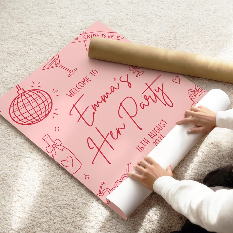 May include: A pink poster with red illustrations and text that reads "Welcome to Emma's Hen Party 16th August 2024". The poster includes drawings of a disco ball, martini glass, and champagne bottle. A rolled-up poster is next to it.