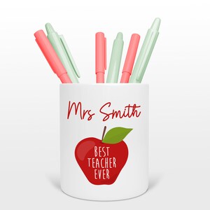 Puede incluir: Porta lápices de cerámica blanca con un gráfico de manzana roja y el texto "Mejor profesor de todos los tiempos". El porta lápices también tiene el nombre "Sra. Smith" escrito en él. Hay seis bolígrafos en el porta lápices, tres son rosas y tres son verde claro.