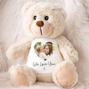 Puede incluir: Un oso de peluche beige con una camiseta blanca con una foto en forma de corazón de una madre y un niño. La camiseta tiene el texto "We Love You" y una pequeña "X" debajo. El oso tiene una nariz marrón y ojos negros.