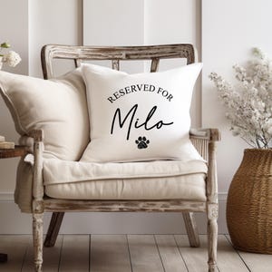 Puede incluir: Un coj&iacute;n blanco con el texto "RESERVED FOR Milo" en escritura negra y una huella de pata negra. El coj&iacute;n est&aacute; sobre una silla color crema con un marco de madera envejecida. Decoraci&oacute;n del hogar.