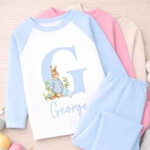 May include: A light blue and white pajama set with a bunny and Easter egg design. The set includes a long-sleeved top with the letter "G" and the name "George" printed on it, and matching blue pants. Other pajama sets are visible.
