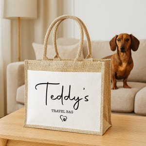 May include: A beige jute tote bag with a white front panel. The text "Teddy's TRAVEL BAG" is written in black script. A brown dachshund dog is in the background.