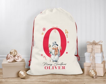 Puede incluir: Una bolsa de regalo de Navidad blanca con una letra O roja, un muñeco de nieve y el texto "Feliz Navidad Oliver".