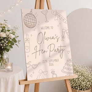 May include: A light beige sign on a wooden easel, featuring the text "Welcome to Olivia's Hen Party" and the date "25th September 2026." The sign is decorated with line drawings of a disco ball, champagne glasses, and a cake.