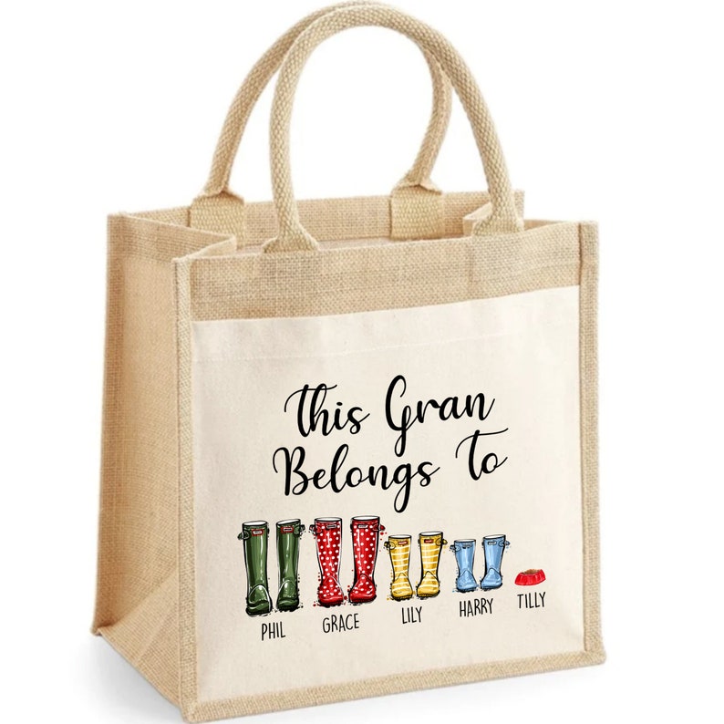 May include: A beige tote bag with a white canvas insert. The bag has a black inscription that reads "This Gran Belongs To" and features a line of colorful rain boots with names underneath: Phil, Grace, Lily, Harry, and Tilly.