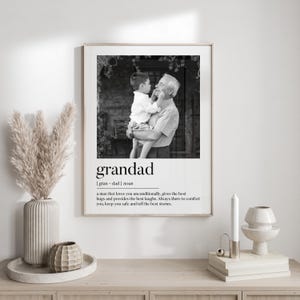 May include: Black and white framed print featuring a photograph of a grandfather holding a child. The word "grandad" is printed below the photo, along with a definition. The print is displayed on a wooden surface.