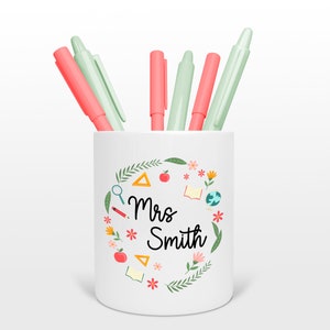 Puede incluir: Porta lápices de cerámica blanca con un diseño floral y el texto "Mrs. Smith". El porta lápices tiene una manzana roja, un lápiz, una lupa, un libro, un globo terráqueo, una regla y flores alrededor del texto. El porta lápices tiene cinco bolígrafos, dos son verde claro y tres son rosa claro.
