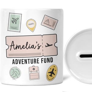 May include: White ceramic piggy bank with a slot on the top. The front of the bank has a pink and white ticket with the text "Amelia's Adventure Fund". The bank is decorated with travel-themed illustrations, including airplanes, stamps, and a globe.