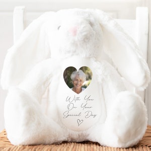 Può includere: Un coniglio di peluche bianco con una finestra a forma di cuore sul petto. La finestra contiene una foto di una donna sorridente. Il coniglio ha il testo "With You On Your Special Day" stampato sul petto.