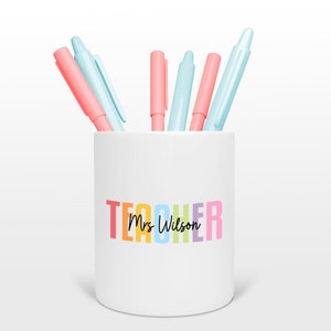 Puede incluir: Porta lápices de cerámica blanca con la palabra "TEACHER" en colores arcoíris y el texto "Mrs. Wilson" en letra cursiva negra. El porta lápices está lleno de bolígrafos rosas y azules.