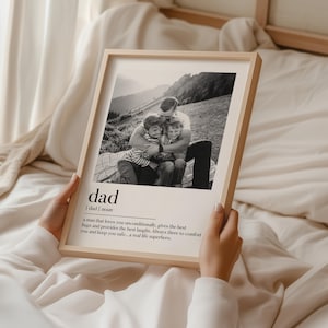 Peut inclure: Un cadre en bois clair contenant une photographie en noir et blanc d'un p&egrave;re &eacute;treignant deux enfants. Le mot "dad" et une d&eacute;finition sont imprim&eacute;s en dessous. L'image est tenue par une personne.