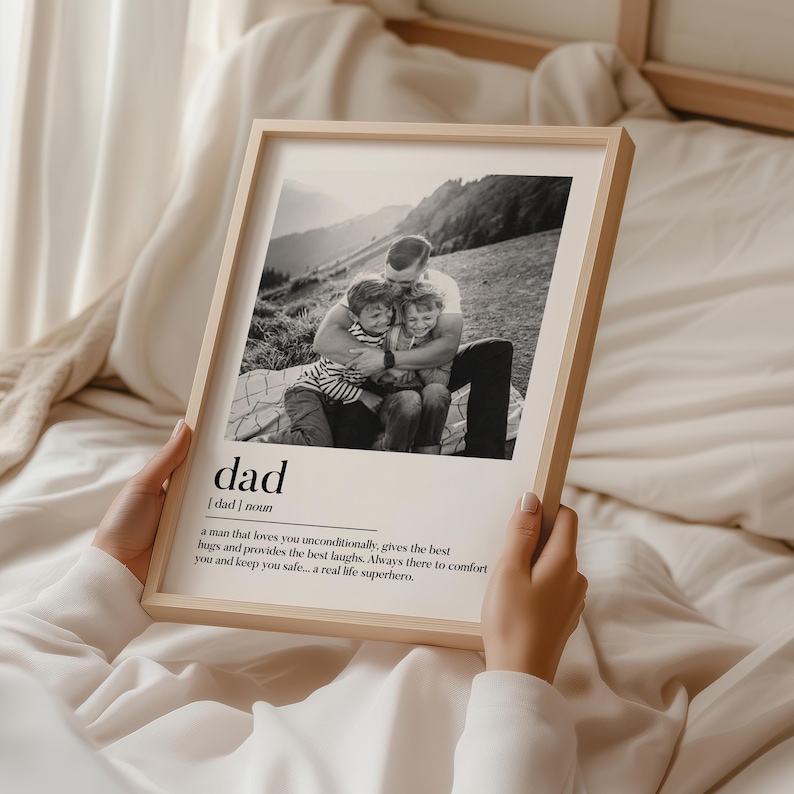 Peut inclure: Un cadre en bois clair contenant une photographie en noir et blanc d'un p&egrave;re &eacute;treignant deux enfants. Le mot "dad" et une d&eacute;finition sont imprim&eacute;s en dessous. L'image est tenue par une personne.