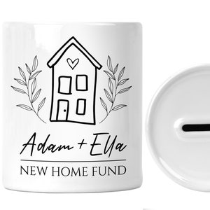 Könnte beinhalten: Weiße Keramik-Sparbüchse mit einer schwarz-weißen Illustration eines Hauses mit einem Herzen über der Tür. Der Text "Adam + Ella NEW HOME FUND" ist unter dem Haus aufgedruckt.