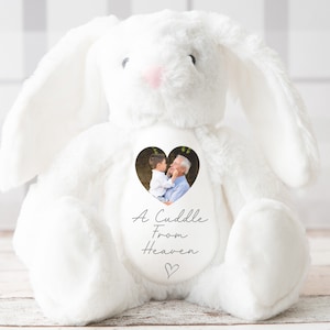 Op de afbeelding: Een witte pluche konijn met een hartvormig raam op zijn borst. Het raam bevat een foto van een jonge jongen en een oudere man. Het konijn heeft de tekst "A Cuddle From Heaven" op zijn borst.