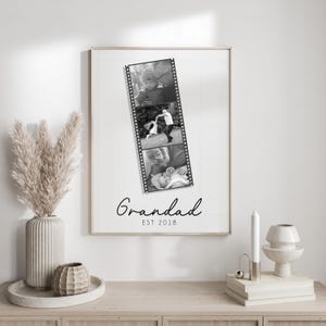 May include: A framed print featuring three black and white photos in a film strip design. The word "Grandad" is written in cursive below the photos, with "EST 2018" underneath. The frame is light-colored.