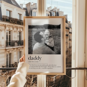 K&ouml;nnte beinhalten: Ein gerahmtes Schwarz-Wei&szlig;-Foto, das einen Vater zeigt, der sein lachendes Kind k&uuml;sst. Der Rahmen ist hellbraun. Unter dem Foto sind das Wort &bdquo;daddy&ldquo; und seine Definition abgedruckt. Im Hintergrund ist ein Fenster mit Blick auf ein Geb&auml;ude zu sehen.