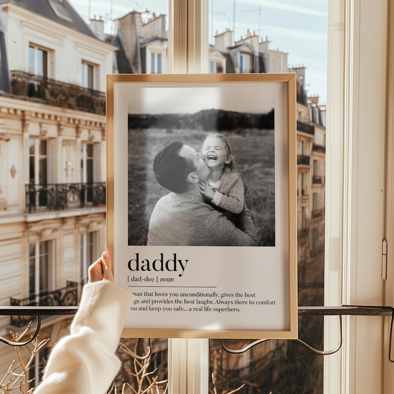 K&ouml;nnte beinhalten: Ein gerahmtes Schwarz-Wei&szlig;-Foto, das einen Vater zeigt, der sein lachendes Kind k&uuml;sst. Der Rahmen ist hellbraun. Unter dem Foto sind das Wort &bdquo;daddy&ldquo; und seine Definition abgedruckt. Im Hintergrund ist ein Fenster mit Blick auf ein Geb&auml;ude zu sehen.