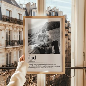 Peut inclure: Un cadre en bois clair contenant une photographie en noir et blanc d'un p&egrave;re &eacute;treignant deux enfants. Le mot "dad" et sa d&eacute;finition sont en dessous. L'arri&egrave;re-plan montre un immeuble et une fen&ecirc;tre.