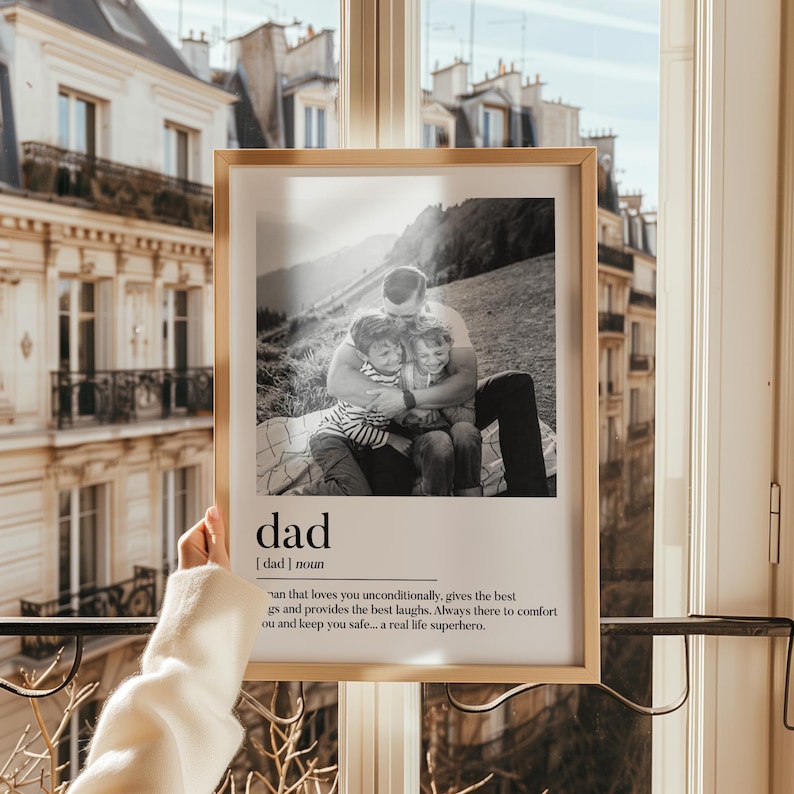 Peut inclure: Un cadre en bois clair contenant une photographie en noir et blanc d'un p&egrave;re &eacute;treignant deux enfants. Le mot "dad" et sa d&eacute;finition sont en dessous. L'arri&egrave;re-plan montre un immeuble et une fen&ecirc;tre.