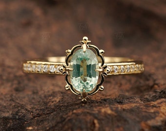 Può includere: Un anello d'oro con una pietra ovale verde circondata da diamanti più piccoli. L'anello ha un design delicato e ornato.