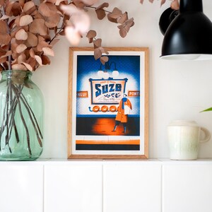 May include: A framed print of a vintage advertisement for "Suze" featuring a woman in a red coat and a blue scarf standing in front of a blue and orange sign with the text "Suze" and "Ap&eacute;ritif d'Orange".