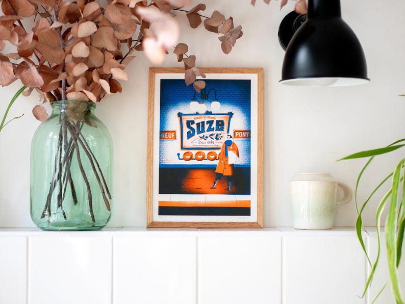 May include: A framed print of a vintage advertisement for "Suze" featuring a woman in a red coat and a blue scarf standing in front of a blue and orange sign with the text "Suze" and "Ap&eacute;ritif d'Orange".