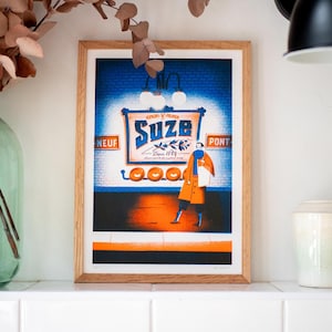 May include: A framed illustration of a woman in a blue coat walking past a sign that reads "Suze" in a blue and white font. The sign is on a brick wall with a blue background. The woman is wearing a blue scarf and carrying a white bag. The sign is lit by a light fixture above it. The illustration is in a wooden frame.