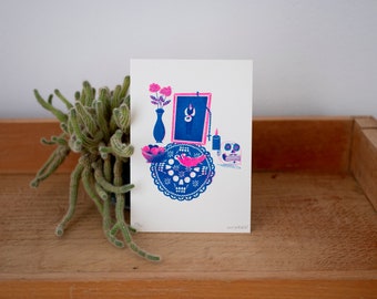 May include: A blue and pink print of a Day of the Dead altar with a skull, flowers, a candle, a vase, and a framed picture.