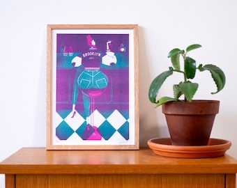 May include: A framed print of a woman in a blue jean jacket and jeans sitting on a bar stool. The print is in shades of pink, blue, and white. The text "BROOKLYN NYC" is visible on the jacket.