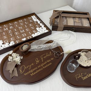 Op de afbeelding: Een set donkerhouten dienbladen en doosjes voor een huwelijksceremonie. Eén dienblad bevat de tekst Derya & Ferhat, een houten ringdoosje, een schaar en witte parels. Een ander dienblad bevat witte snoepjes en een doosje met de tekst Allahin emriyle.