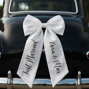 Könnte beinhalten: Eine große weiße Schleife schmückt das Heck eines schwarzen Oldtimers. Die Schleife hat lange Bänder mit den Worten "Just Married" und "Esra & Max" in eleganter Schrift. Der verchromte Stoßfänger des Autos glänzt.