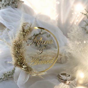 Pode incluir: Um sinal circular de acrílico dourado com os nomes "Dilara & Ahmed" escritos em cursiva. O sinal é decorado com grama de pampas seca e respiração de bebê.