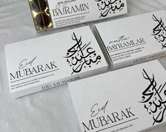 Può includere: Scatole bianche con calligrafia nera e le parole "Eid Mubarak" e "Gezuar Bajramin". Alcune scatole hanno testo in inglese e turco. Una scatola contiene tre dolcetti rotondi marrone scuro.