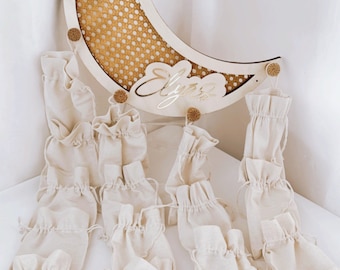 May include: A wooden crescent moon with a woven pattern and the name "Elyse" written on it.  The moon is hanging from a string of small, white fabric pouches.